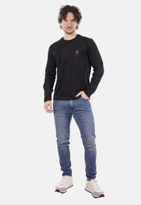 Maglietta nera a maniche lunghe con un logo discreto sul petto, abbinata a jeans blu e sneakers grigie, in piedi su uno sfondo bianco.