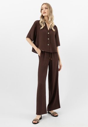 Vrouw met lang blond haar die een losbruine blouse met knopen draagt, bijpassende wijduitlopende broek en zwarte sandalen met open teen en een geweven patroon.