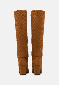 Bottes marron matelassées montant jusqu'aux genoux, avec talons carrés et une sangle à boucle dorée à l'arrière.