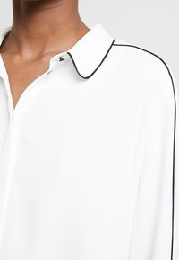 Primo piano di una persona che indossa una blusa bianca con rifiniture nere lungo il colletto e la cucitura della spalla.