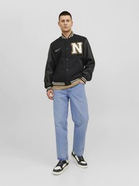 Veste universitaire noire avec logo « N » doré, poignets rayés et col côtelé, portée sur une chemise beige, associée à un jean bleu clair et des baskets.