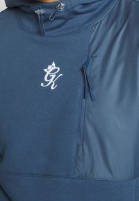 Felpa con cappuccio blu navy, dotata di tasca sul petto con zip, tessuto bicolore e logo bianco a contrasto. Presenta un cappuccio con cordino regolabile.