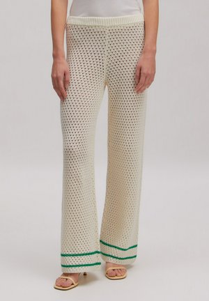 Cremefarbene Häkelhose mit grünen Streifen nahe am Saum, getragen mit beigen Sandalen mit offenen Zehen und einem weißen Oberteil.