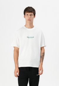 T-shirt bianco in cotone a maniche corte, scollatura rotonda e testo verde "HIDDEN BUNGALOW" sul petto. Indossato con pantaloni jogger neri.