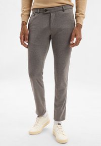 Pantaloni grigi su misura con una texture liscia, dotati di una piega frontale, tasche laterali e chiusura con bottone, abbinati a sneakers bianche.