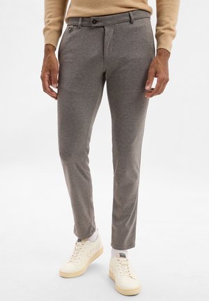 Pantaloni grigi su misura con una texture liscia, dotati di una piega frontale, tasche laterali e chiusura con bottone, abbinati a sneakers bianche.