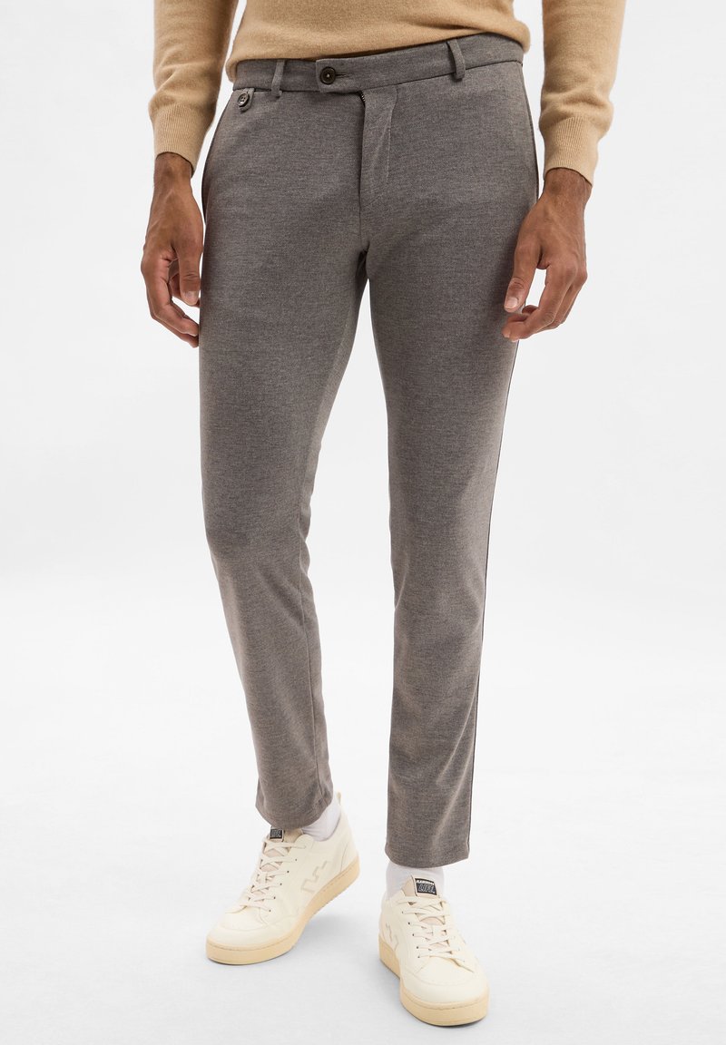 Pantaloni grigi su misura con una texture liscia, dotati di una piega frontale, tasche laterali e chiusura con bottone, abbinati a sneakers bianche.