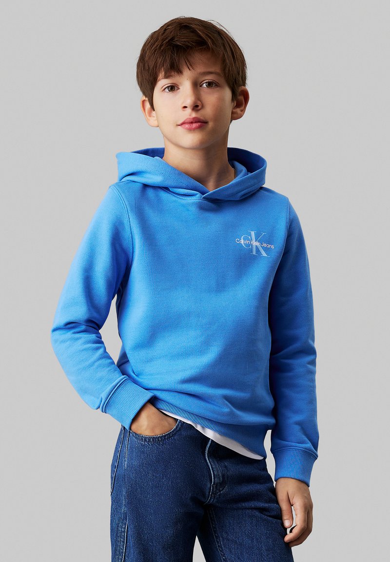 Sudadera con capucha azul hecha de suave tela de algodón, con un logo en la parte frontal y puños acanalados. Combinada con jeans de mezclilla oscura.