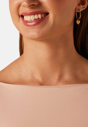 Femme souriante, portant des boucles d'oreilles créoles en or avec des breloques en forme de cœur, et un haut rose clair à épaules dénudées, sur un fond uni.