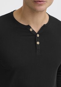 Schwarzes Langarm-Henley-Shirt aus weichem Stoff. Verfügt über einen runden Ausschnitt und drei Holzknöpfe an der Blende. Einfaches Design.