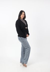Schwarze gestrickte Strickjacke mit tiefem V-Ausschnitt, ausgestattet mit einem goldenen Knopf. Kombiniert mit hellblauen, hochtaillierten Jeans und klaren, absatzlosen Sandalen.