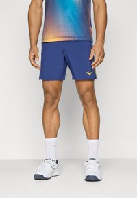 Blaue Sportshorts mit einem Logo am linken Bein. Modell trägt weiße Schuhe und Socken. Das Oberteil hat ein Farbverlaufdesign in Blau und Orange.