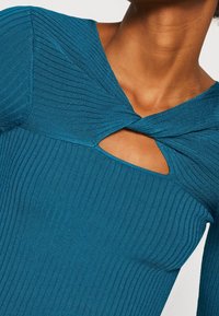 Vihreäsininen ribbineulottu yläosa, jossa on kiertynyt design pääntiessä ja avainreikänauha. Kangas on sileäpintainen ja istuva.