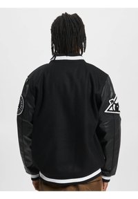 Veste de varsity noire avec un corps en laine et des manches en cuir. Présente un col et des poignets à rayures blanches, ainsi que des patchs brodés sur les deux manches.