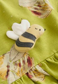 Petit jouet en peluche en forme d'abeille avec des rayures noires et des ailes blanches cousues sur un tissu vert à motifs floraux.