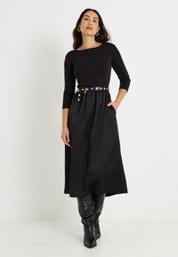 Vestido negro de manga larga con un cinturón que presenta acentos circulares dorados, fabricado con una tela suave. Modelo con botas negras hasta la rodilla.