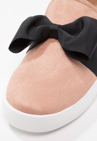 Chaussure en suède rose pâle avec un large nœud en tissu côtelé noir sur le dessus et une semelle en caoutchouc blanche.