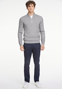 Maglione grigio chiaro con zip e collo alto realizzato in un materiale morbido, abbinato a pantaloni blu navy. Il modello è in piedi su uno sfondo bianco e indossa sneakers bianche.