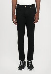 Μαύρα τζιν παντελόνια με στενή γραμμή, επιδιορθωμένα με χρυσές λεπτομέρειες, πλάγιες και πίσω τσέπες, και λεία υφή. Φοριούνται με μαύρα αθλητικά παπούτσια.