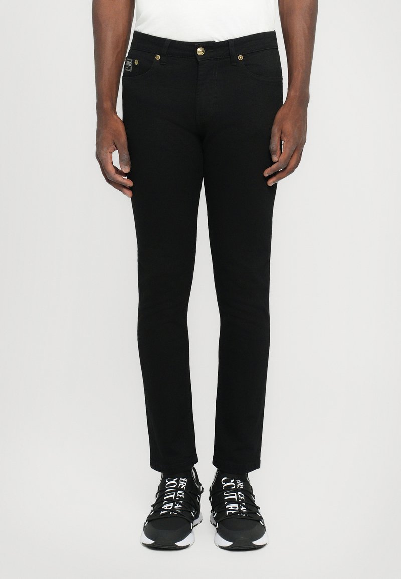 Μαύρα τζιν παντελόνια με στενή γραμμή, επιδιορθωμένα με χρυσές λεπτομέρειες, πλάγιες και πίσω τσέπες, και λεία υφή. Φοριούνται με μαύρα αθλητικά παπούτσια.