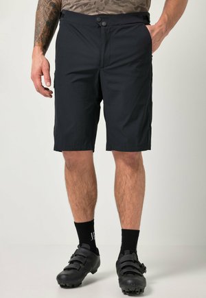 Mann trägt schwarze knielange Shorts, schwarze Socken und schwarze Fahrradschuhe und steht vor einem schlichten Hintergrund.