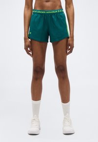 Αθλητής που φοράει τυρκουάζ σορτς Under Armour με κίτρινες λεπτομέρειες, λευκές κάλτσες και λευκά αθλητικά παπούτσια, στέκεται μπροστά σε απλό φόντο.