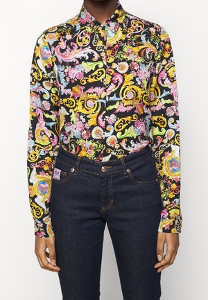 Kobieta ubrana w czarną bluzkę z długim rękawem, zdobioną kolorowymi kwiatami i barokowymi zawijasami, w połączeniu z ciemnoniebieskimi dżinsami.