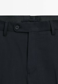 Donkerblauwe broek van een gestructureerde stof, met een knoopsluiting, plooien aan de voorkant en riemlussen. Geen zakken zichtbaar vanuit deze hoek.