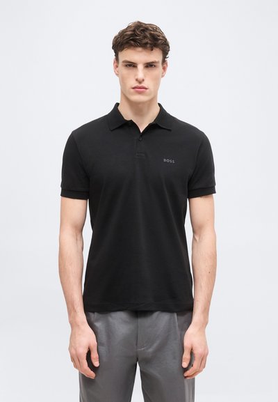 Homme portant un polo noir BOSS à manches courtes et un pantalon gris, debout devant un fond clair uni, faisant face à l'avant.