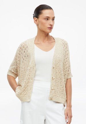 Vrouw draagt een beige gebreid vest met korte mouwen en knopen over een witte top en witte broek, staand met één hand in de zak.