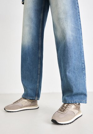 Beige Sport-Schuhe mit Mesh- und Lederpaneelen, ausgestattet mit einer schwarzen Außensohle und Schnürdesign, kombiniert mit hellblauen, lockeren Jeans.