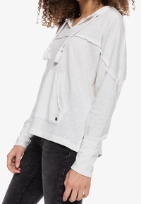 Sudadera blanca con capucha y acabado texturizado, cierre con cordón, bolsillo frontal y aberturas laterales. Mangas largas con puños enrollados.
