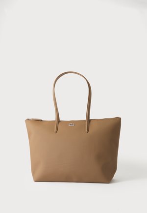 Sac fourre-tout beige avec deux poignées et un petit emblème logo argenté, sur un fond blanc uni.