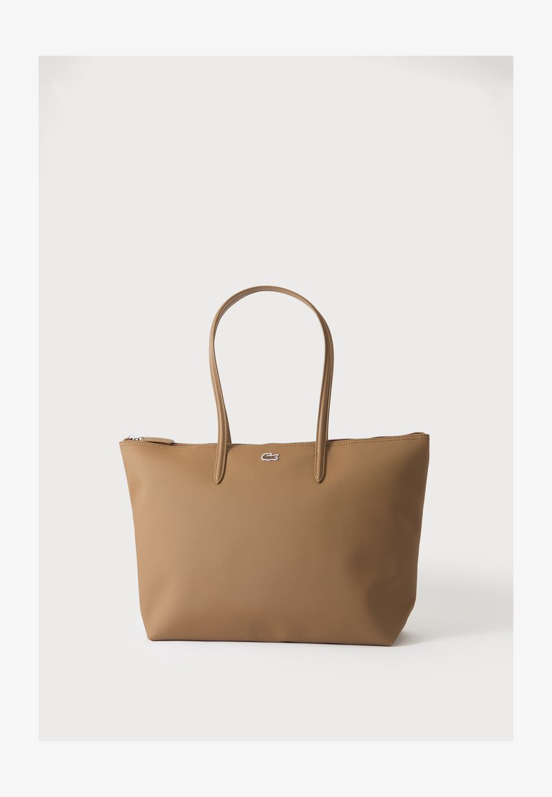 Sac fourre-tout beige avec deux poignées et un petit emblème logo argenté, sur un fond blanc uni.