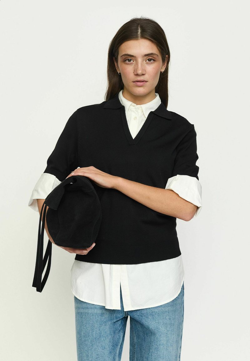 Femme aux cheveux bruns et lisses porte un pull noir sur une chemise blanche, un jean bleu, tient un sac noir, debout sur un fond uni.