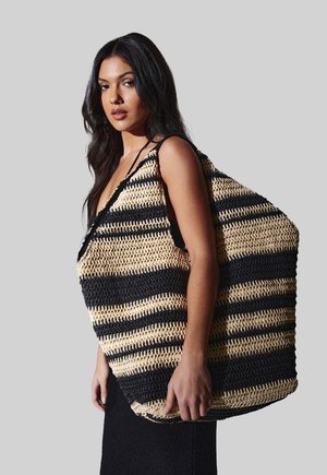 Mujer con cabello largo y oscuro lleva un gran bolso tote tejido a rayas negras y beige sobre el hombro, con fondo gris.