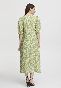 Robe midi florale verte avec manches bouffantes, poignets élastiques et fermeture éclair au dos. Talons noirs à lanières visibles en bas.