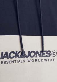 Sudadera en bloques de color negro y beige con cordones, que presenta un logotipo audaz en texto gris oscuro en la parte frontal, fabricada en material de algodón.