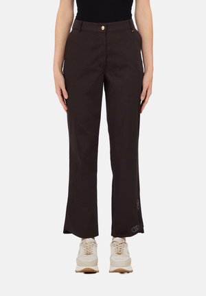 Donna che indossa pantaloni marrone scuro a gamba dritta con bottone dorato e sneakers bianche, in piedi contro uno sfondo semplice.