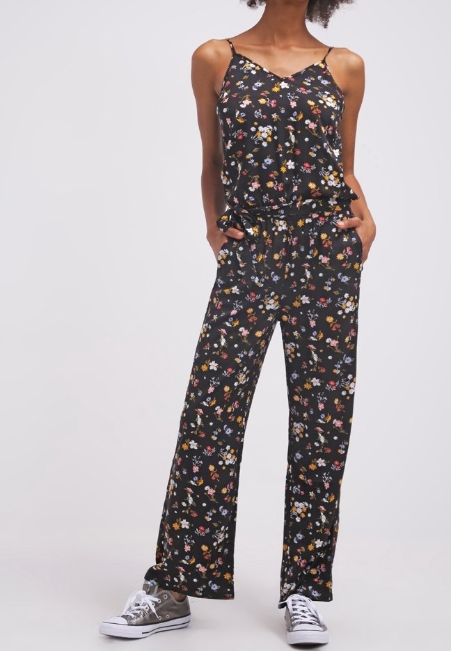 Femme portant une combinaison sans manches noire à motifs floraux, jambes larges, et des baskets grises, debout avec les mains dans les poches devant un fond uni.