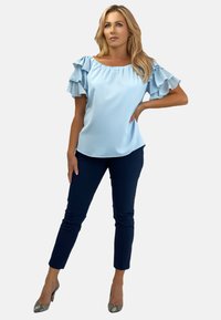 Jasnoniebieska bluzka odkrywająca ramiona z falbaniastymi rękawami; połączona z granatowymi dopasowanymi spodniami i wzorzystymi szpilkami. Gładki materiał, elegancki design.