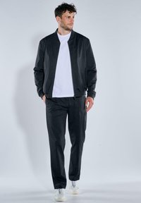 Schwarze Bomberjacke mit glatter Textur, kombiniert mit passenden Hosen, über einem weißen T-Shirt getragen. Weiße Sneaker runden das Outfit ab.