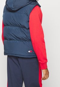 Granatowa pikowana kamizelka z kapturem oraz czerwona koszula z długim rękawem pod spodem. Na dole kamizelki znajduje się mała naszywka z logo.