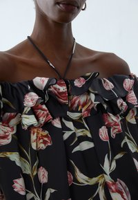 Off-the-shoulder blouse featuring a black floral pattern with ruffled accents, made of lightweight fabric. Key colors include pink and green.