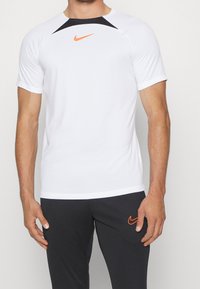 Biała koszulka sportowa z krótkim rękawem, czarne akcenty na ramionach i pomarańczowe logo Nike. Gładki materiał o dopasowanym kroju.