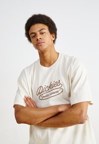 Kreminės spalvos marškiniai su rudo lyno tipo logotipu, kuris sako "Dickies Quality Garments." Trumpi rankovės, laisvas kirpimas, medvilnės medžiaga.