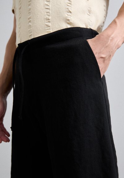 Sorte bukser med en struktureret overflade og elastisk talje. Har sidelommer og en løs pasform. Styliseret med en lysfarvet top.