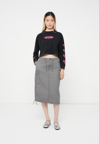 Tânără îmbrăcată cu un hanorac cropped negru cu text roz, fustă midi cargo gri și pantofi Mary Jane argintii, stând sprijinită de un zid alb.