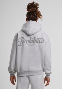 Pilka sportinė striukė su dideliu užrašu "Draudžiama" ant nugaros, turinti raištelių gobtuvą ir įmantrius rankogalius. Minkšta medžiaga ir laisvas pjūvis.