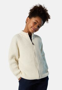 Krämfarbener gerippter Pullover mit hohem Kragen und Reißverschluss, mit langen Ärmeln und seitlichen Taschen, über einem schwarzen Hemd getragen.
